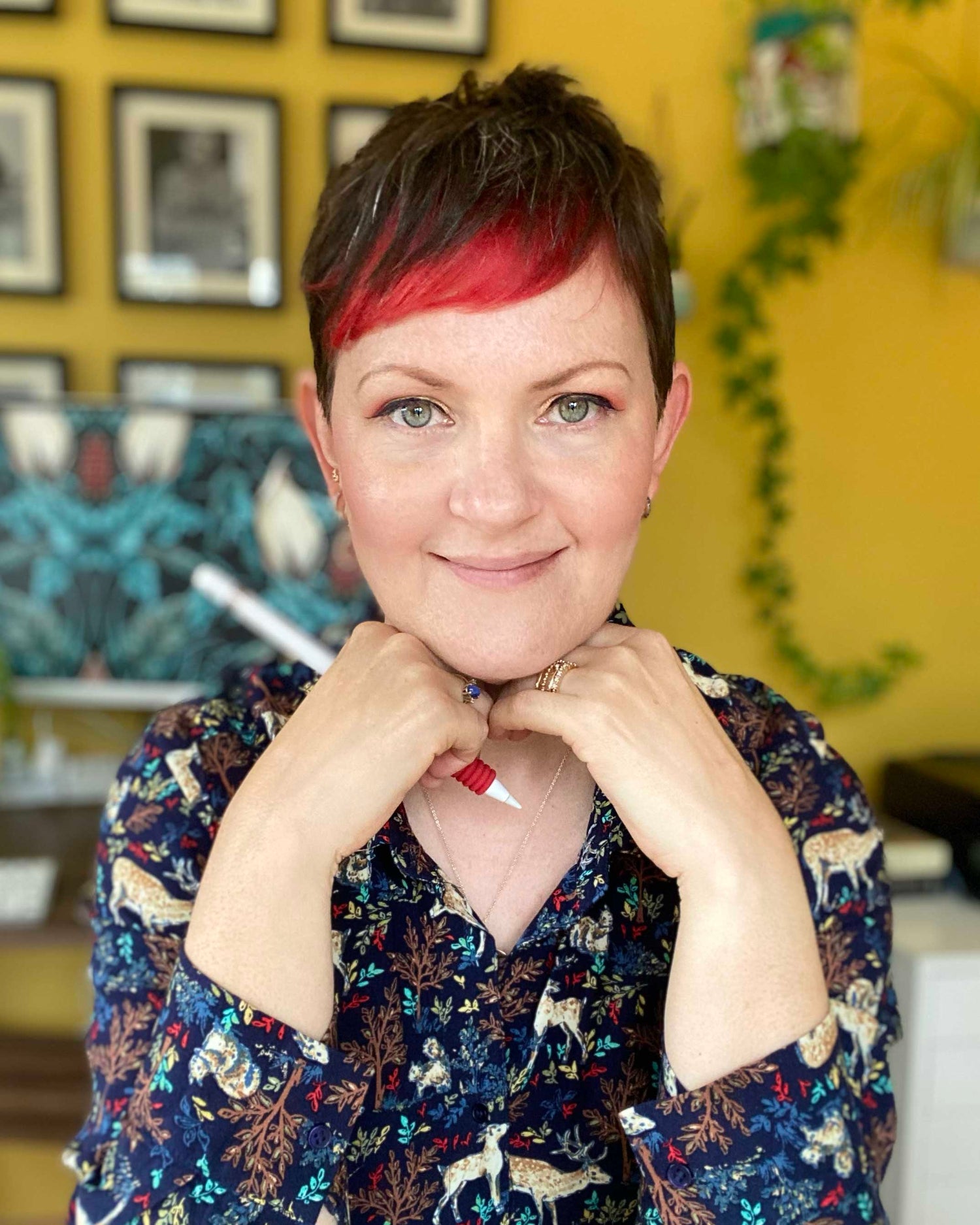 April Mawhinney in a shirt with woodland creatures, holding apple pencil and sitting in front of a yellow wall in her studio.