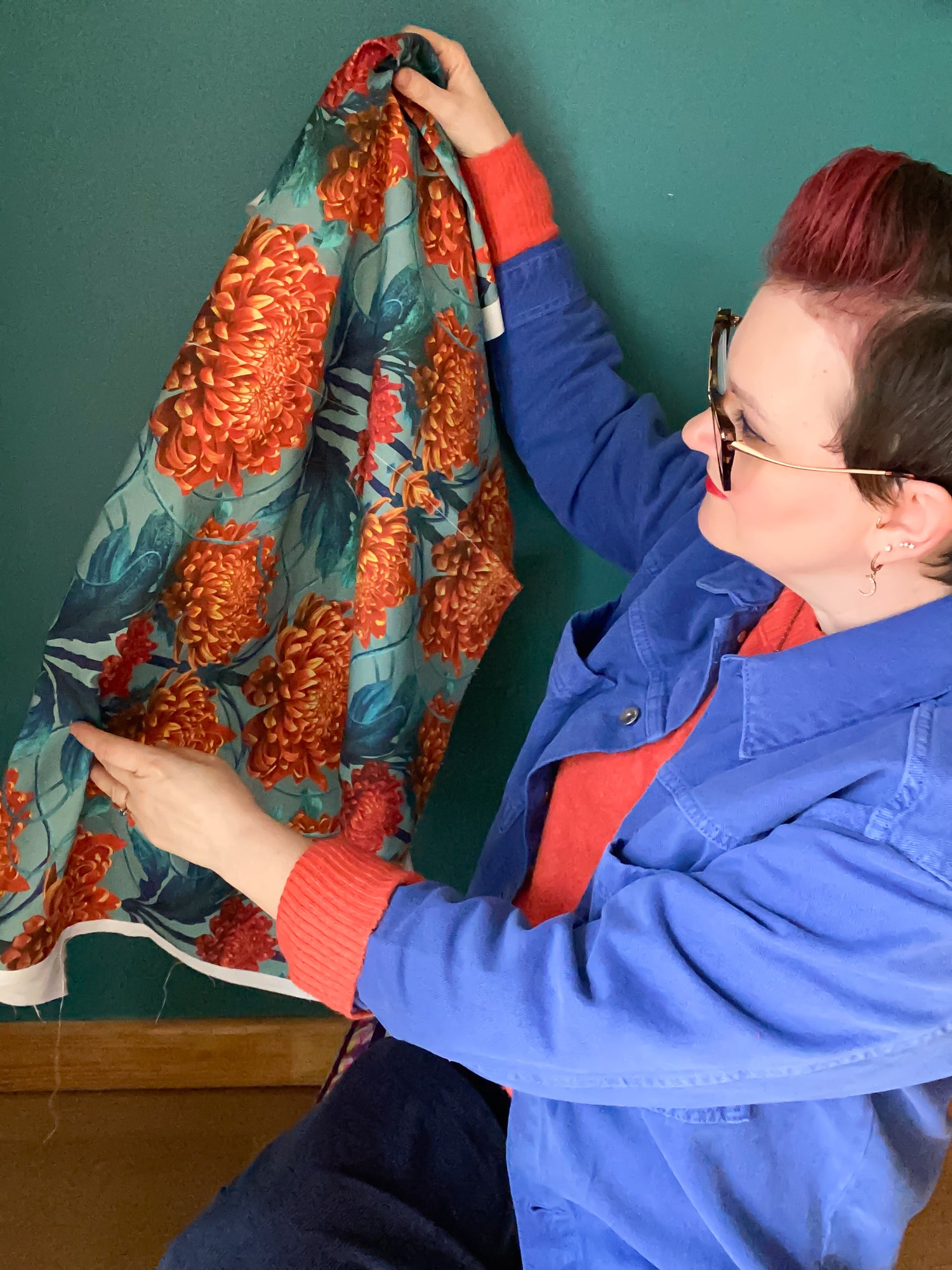 Textile and wallpaper design, April Mawhinney, holding up fabric printed with her Caustic Chrysanthemum in Orpiment Orange design. She is in front of a teal wall. April is wearing a indigo chore jacket, with an orange jumper. She has a red quiff hairstyle are large quirky horn-rimmed glasses