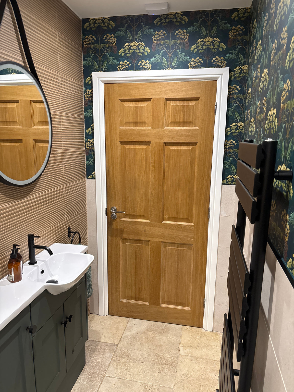Bathroom, Hemlock Wallpaper in Bold Brassica, UK. A six-panel wooden bathroom door with a silver handle, set in a white frame within a bathroom featuring dark floral wallpaper, a stone-tiled wall, a white sink, and a black heated towel rail.