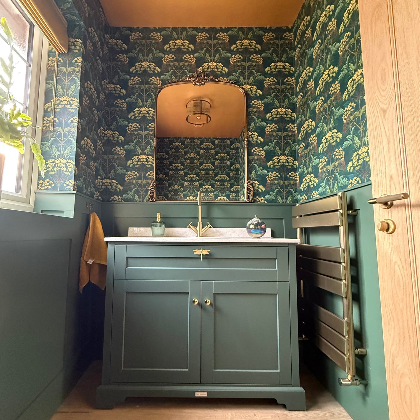 Customer's half bathroom/cloakroom decorated with green and yellow Hemlock Wallpaper in colourway Bold Brassica. The wooden floor and wooden door are a pael pine colour and all the metal finishes are brushed brass. There is a green painted wooden vanity unit topped with marble and a large vintage Rocco style mirror about the sink. Ceiling is painted a dark ochre yellow.