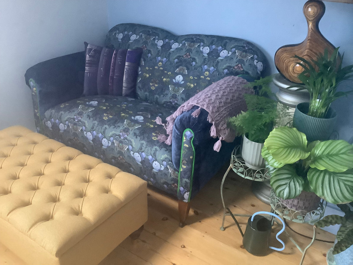 Home Office, A vintage sofa upholstered in Bloomsbury Moth Linen in a cozy room setting, styled with purple pillows, a pink knit throw blanket, a yellow tufted ottoman, and potted plants on metal stands.