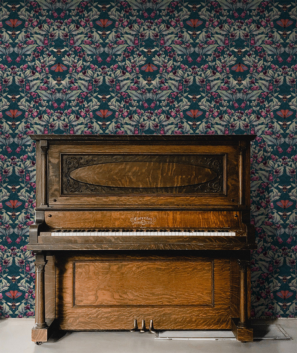 a dark wood upright piano with the word 'Clarendon' on the front sits in fron of a wall wallpapered in April Mawhinney's dark blue version of the Deadly Nightshade wallpaper called Midnight. It show muted green and blue foliage, purple berries and magenta flowers with bees, and red and purple cinnabar moths.