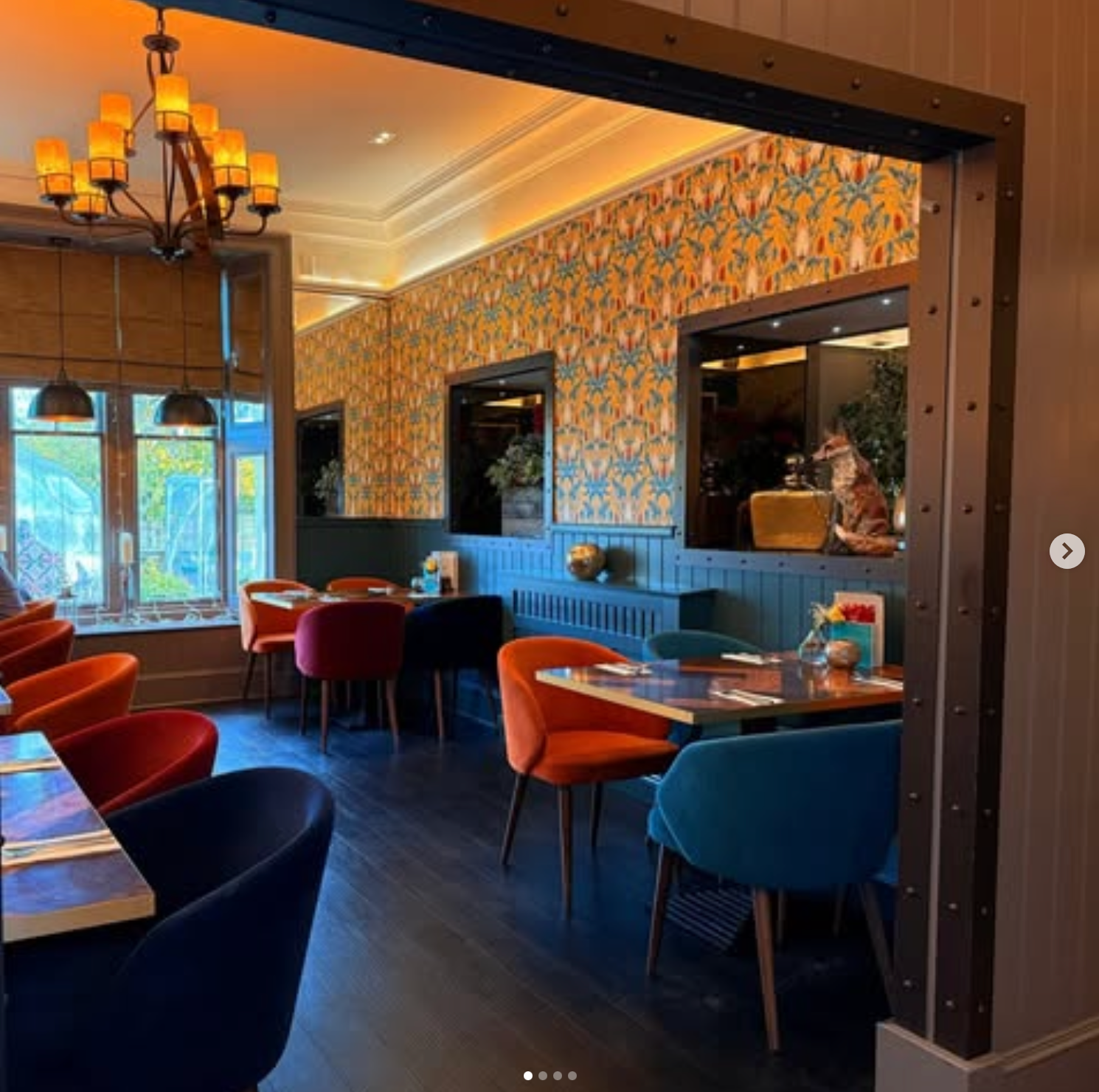 Dining area of the The Fox & Willow, Ayr, Scotland, featuring April Mawhinney's Lords and Ladies wallpaper in Alchemist's Yellow. There is dark blue woodwork, dark brown wooden floor and dining tables with velvet seats in Magenta, orange and navy.