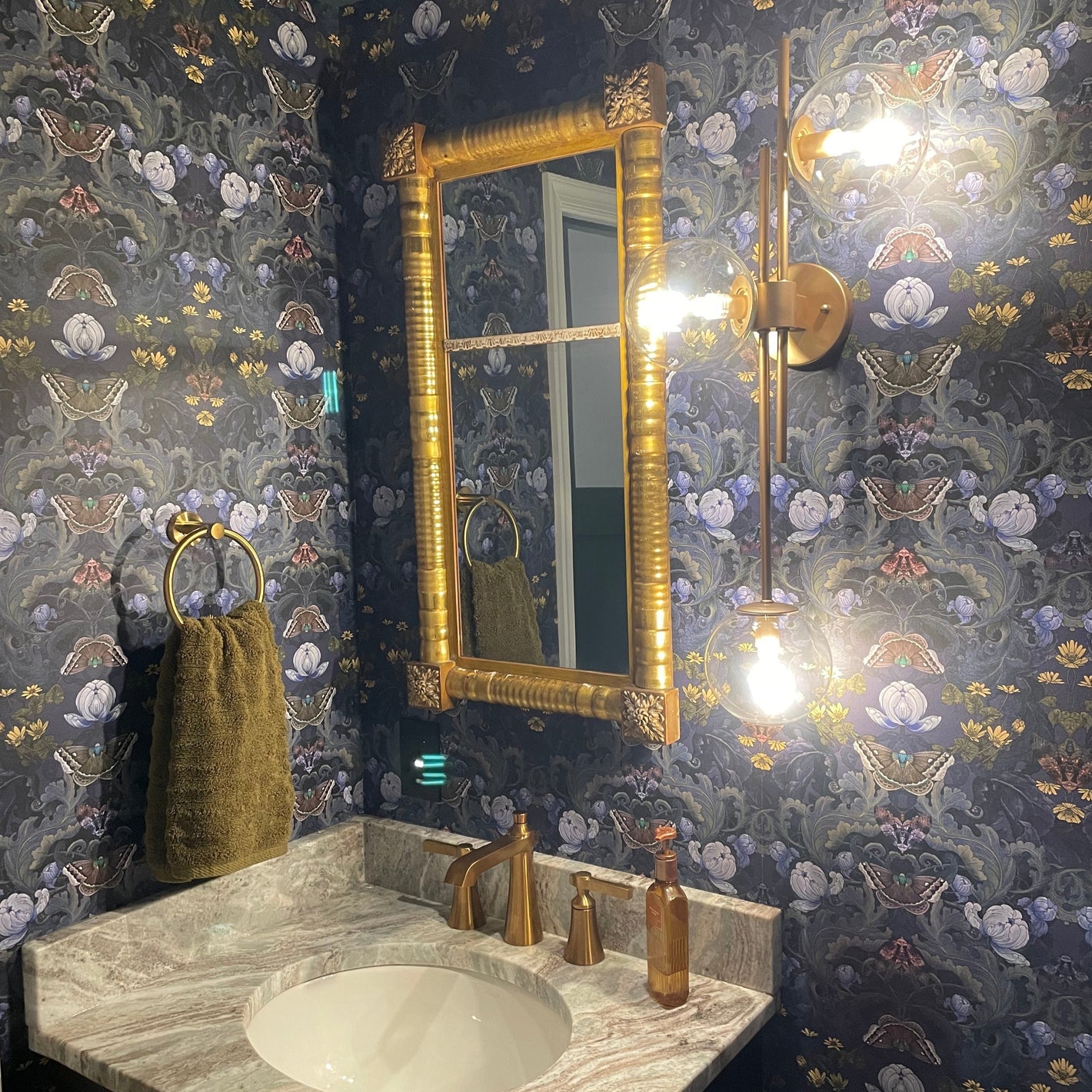 A bathroom with April Mawhinney's dark and moody Bloomsbury Moth Wallpaper. The sink is topped with marble and the taps, towel rail and wall sconce are brushed brass. There is a large vintage rectangular mirror above the sink. 