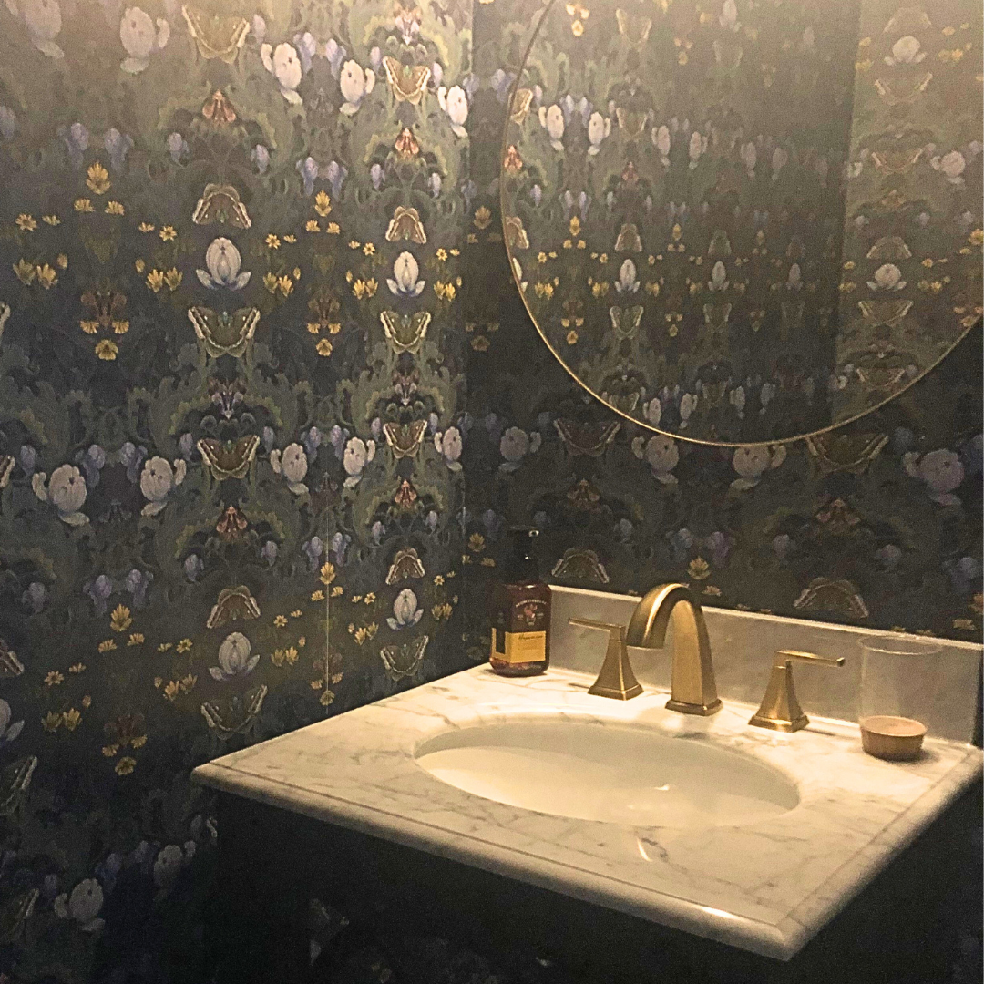customer cloakroom/half-bath featuring dark William Morris inspired 'Bloomsbury Moth' wallpaper. All walls are wallpapered and it created a dark luxurious space. There is a large circular mirror on the wall. The vanity has a marble top and the facet/taps are in brushed brass.