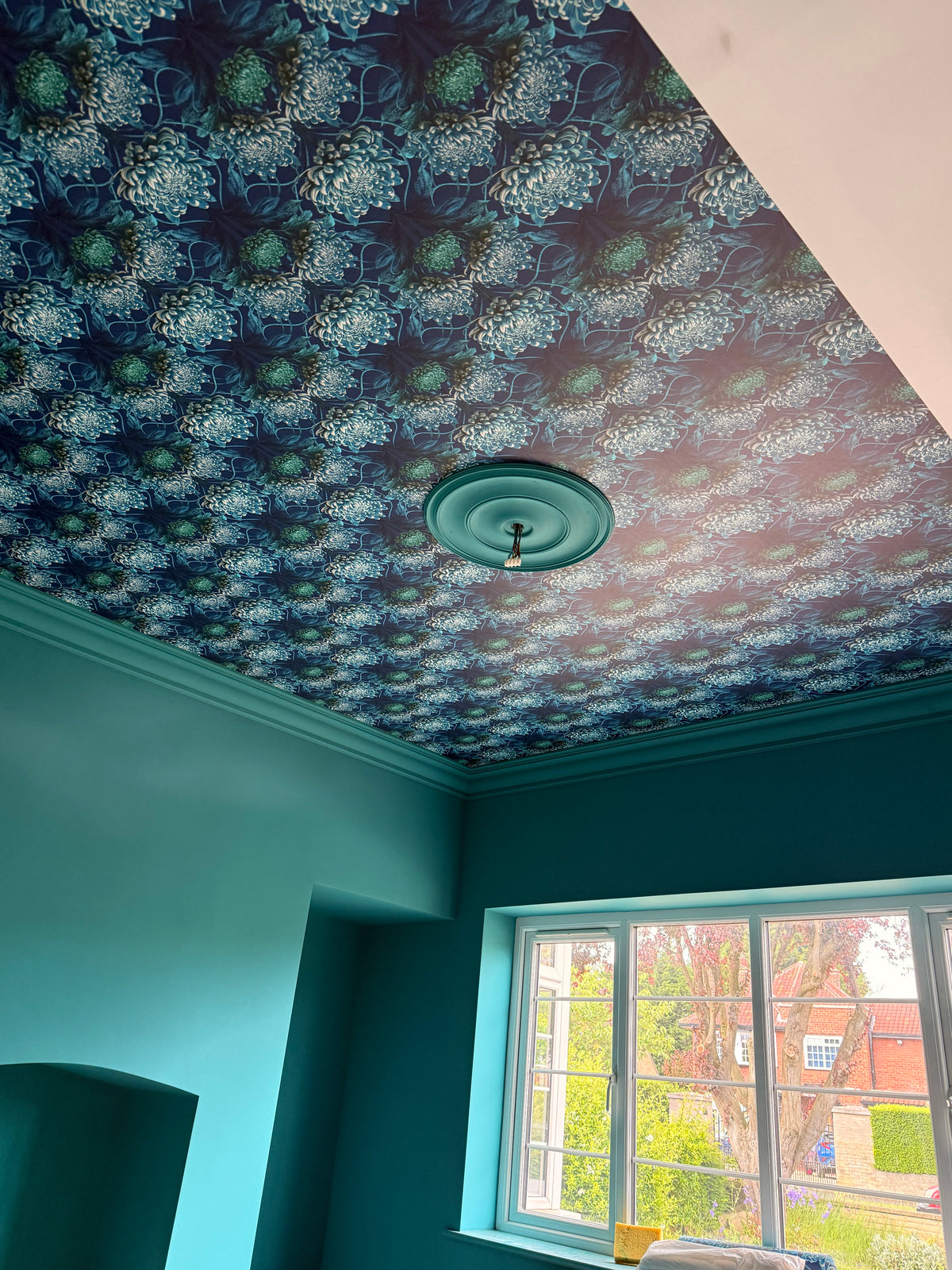 A room corner showing Wallpapered Ceiling, Caustic Chrysanthemum in Blue Vitriol, UK. Teal painted walls, with a window and outdoor view visible.
