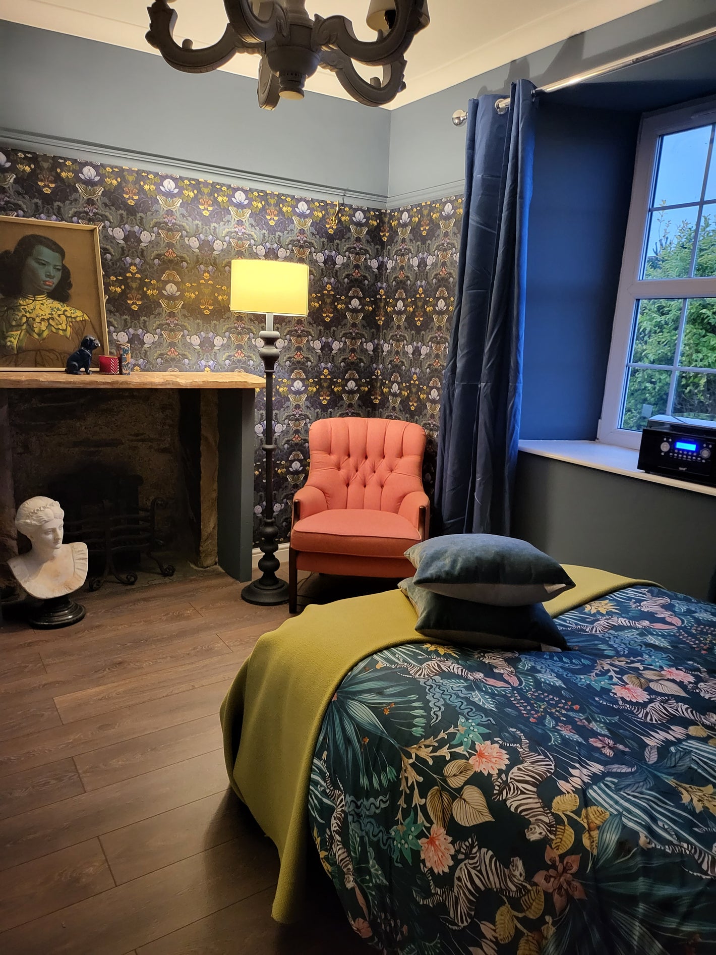 Beautiful bedroom with April Mawhinney's Bloomsbury Moth wallpaper on the walls. The wallpaper is coloured in navy blues, greens and browns, in a swirling William Morris inspired design. It has moths and yellow celadine flowers on it. The bedroom has blue velvet curtains, and a coral linen button backed armchair in the corner. There is a large standard lamp, a bed with a navy tropical inspired duvet cover and a mustard yellow blanket.